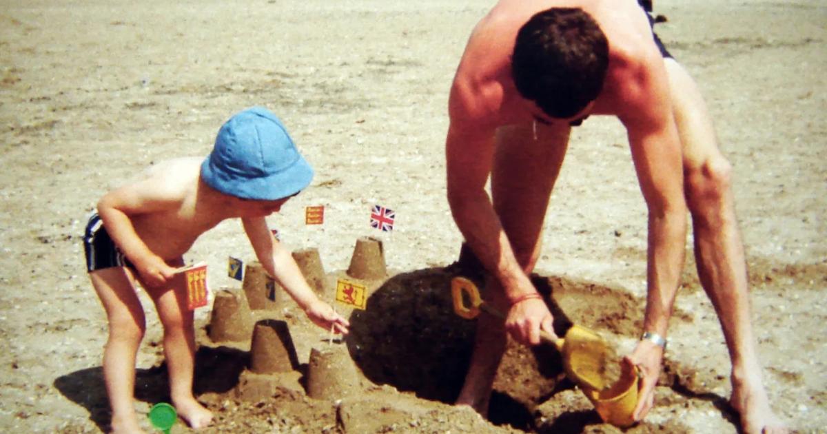 A colour photograph of a man and a small child in a sun hat building and digging at sandcastles on a wide flat beach.