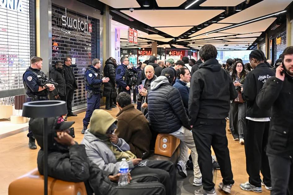 A crowd waiting outside a Swatch store in a shopping arcade, some sitting on the floor with bags, uniformed police standing among them.