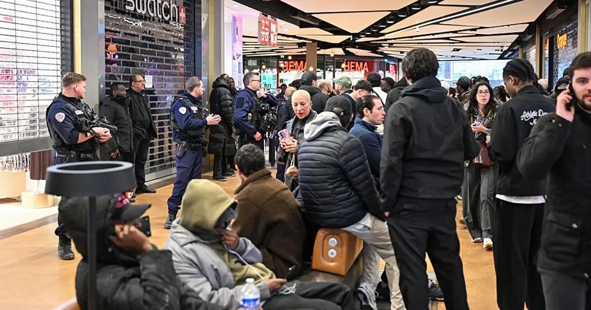 A crowd waiting outside a Swatch store in a shopping arcade, some sitting on the floor with bags, uniformed police standing among them.