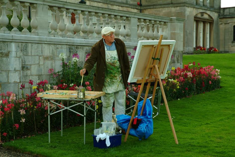 An older man in an apron and cap painting at an easel on a lawn beside a stone balustrade, brushes and pots on a folding table and flowerbeds behind him.