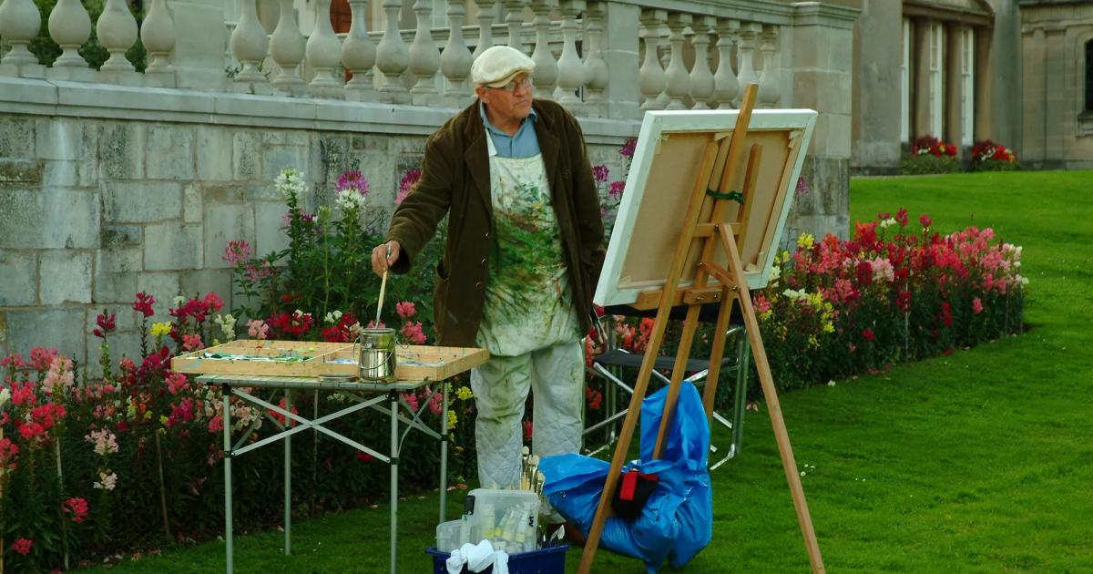 An older man in an apron and cap painting at an easel on a lawn beside a stone balustrade, brushes and pots on a folding table and flowerbeds behind him.