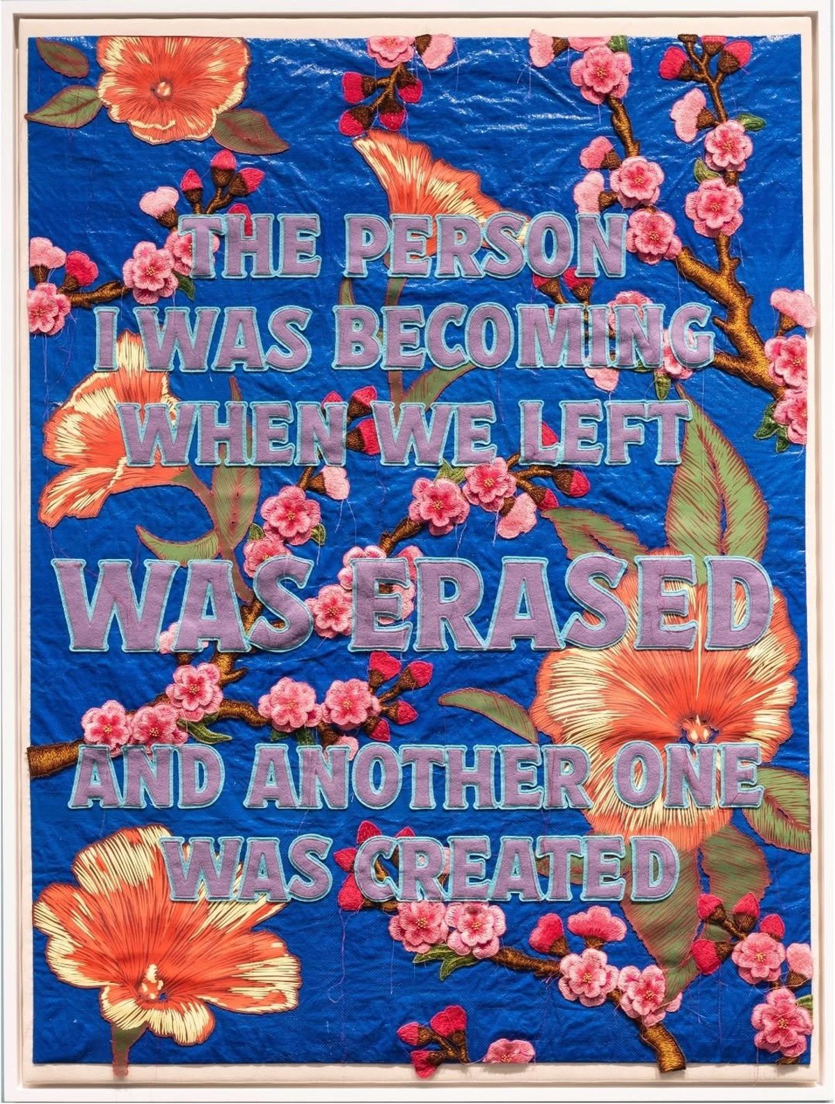 A framed embroidered banner on blue ground reading, the person I was becoming when we left was erased and another one was created, the lettering surrounded by appliqued blossom and orange flowers.