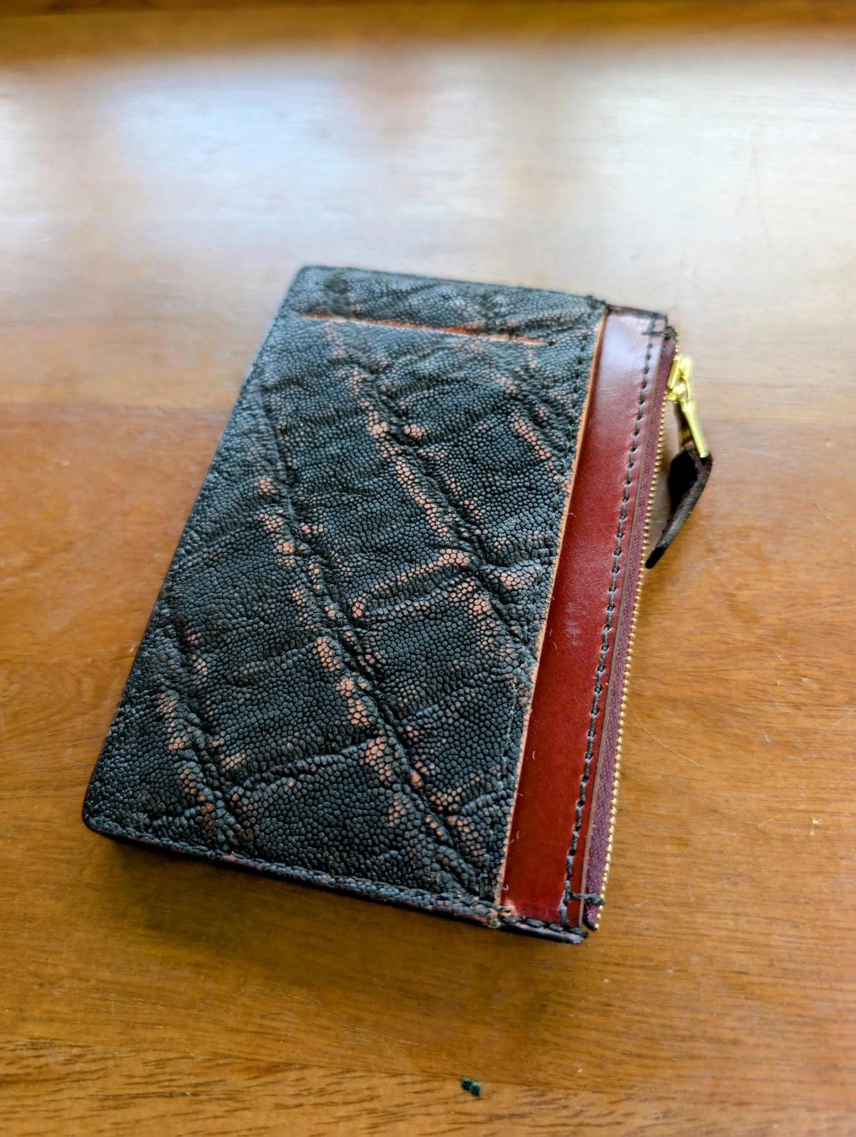 A slim wallet on a wooden table, its front panel in dark stitched textured leather with a tan leather spine and a brass zip pull.