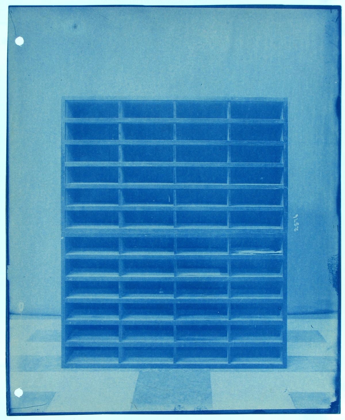 A cyanotype photograph of a tall wooden cabinet of forty eight empty rectangular compartments, standing against a plain wall.