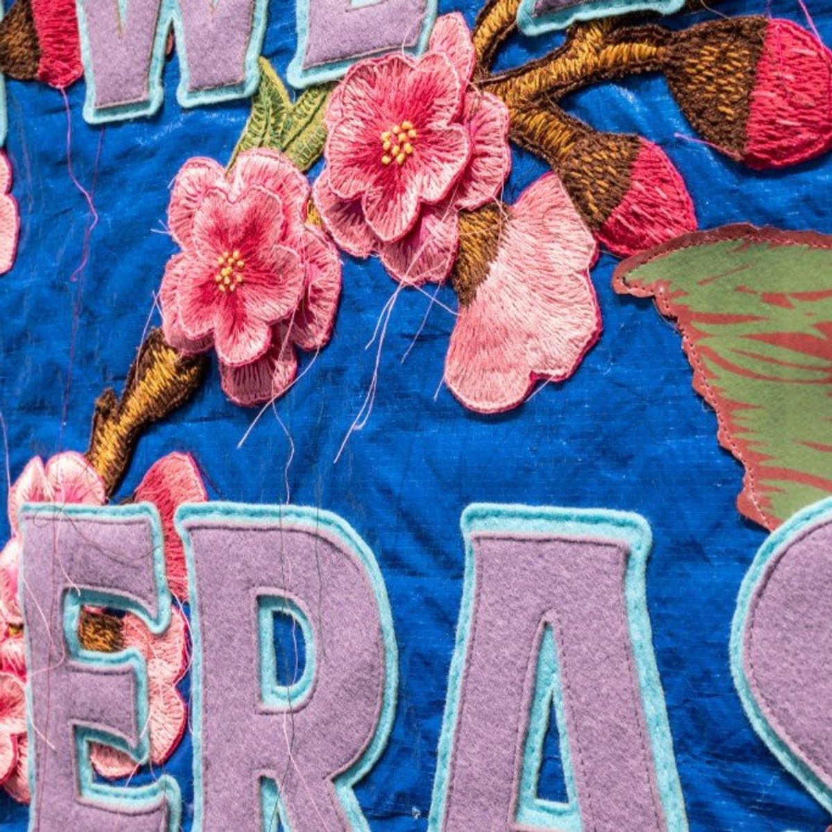 A closer detail of the same banner, the word erased spelled in lilac appliqued letters among heavily stitched pink flowers and loose threads.