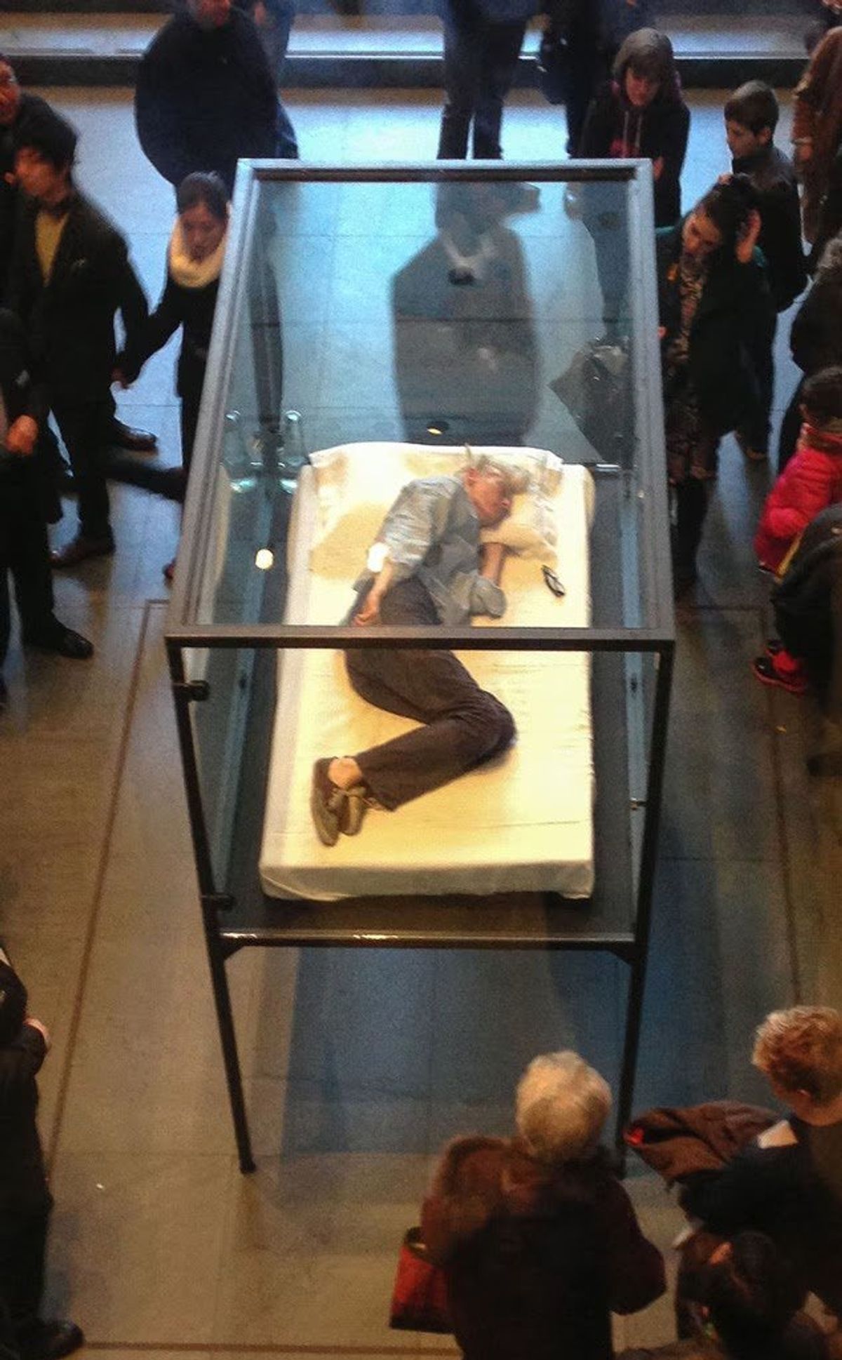 A crowd standing around a glass vitrine in a gallery containing a person lying on a mattress inside it.