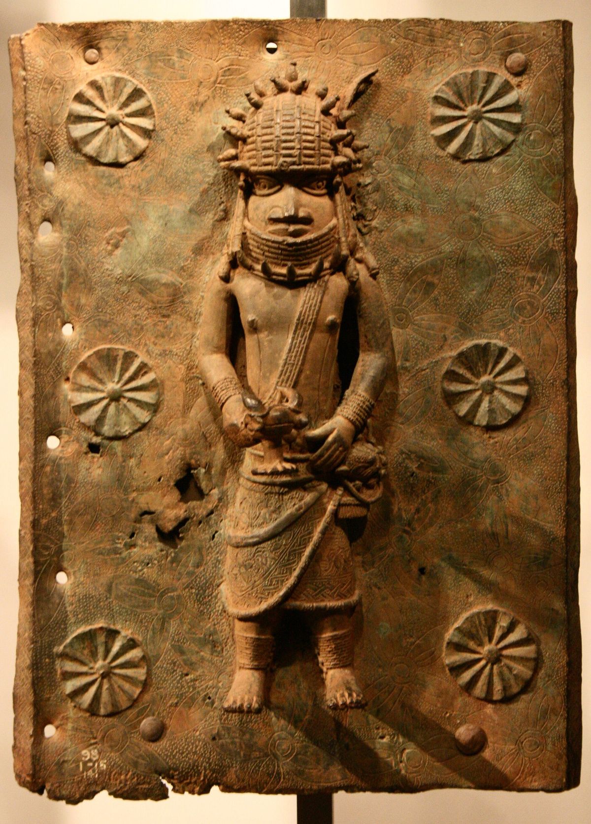 A rectangular Benin brass plaque in relief showing a standing figure in a beaded collar and headdress, with rosettes across the background and an accession number stamped at the lower left.