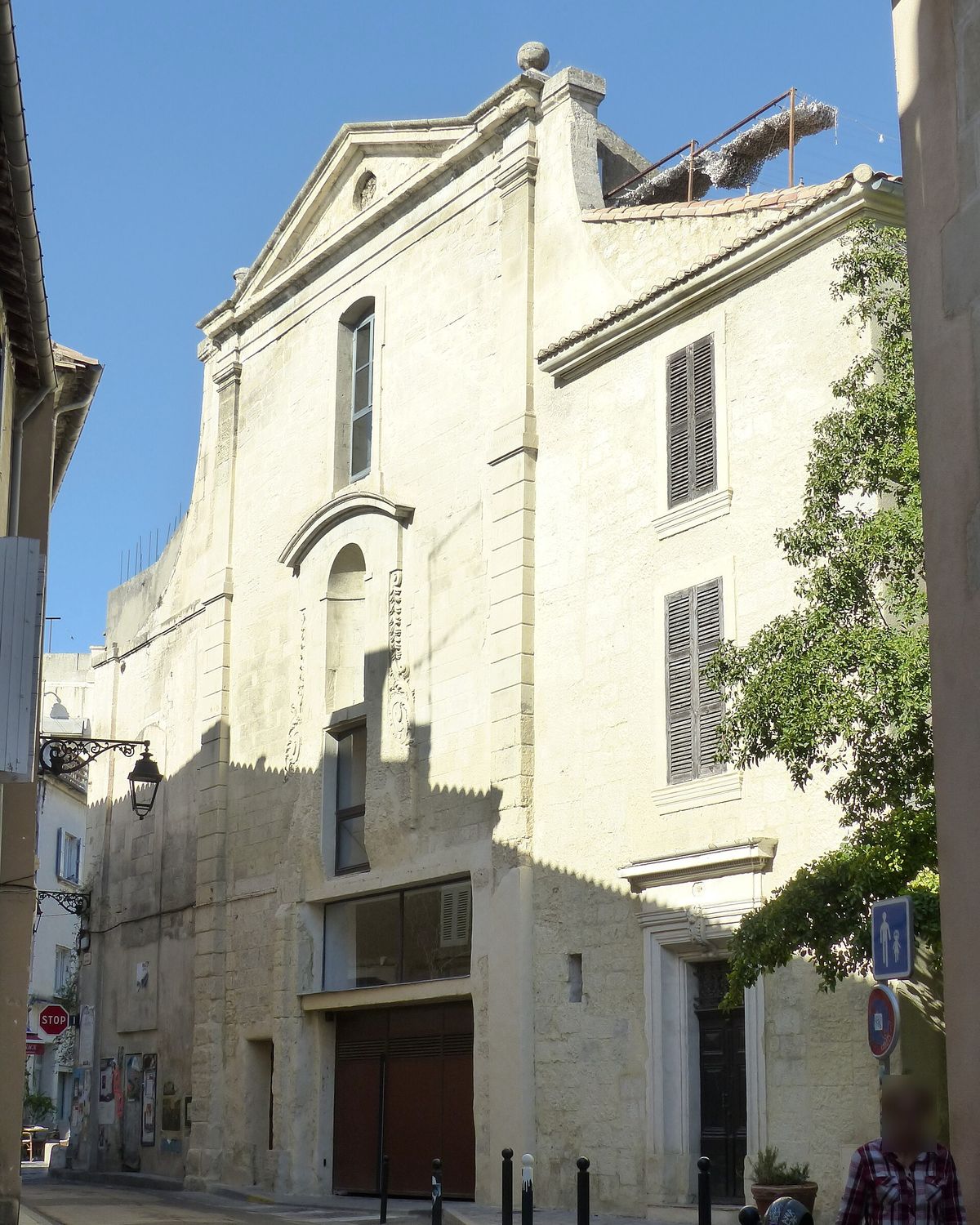 The former Sainte-Croix church in Arles seen from the street: a pale stone building with a pedimented gable, shuttered windows and closed red doors.