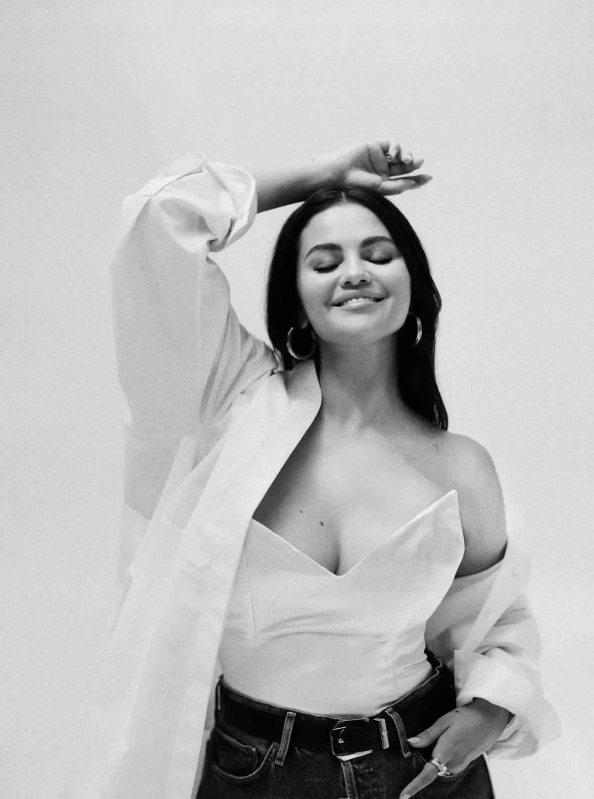 A black and white portrait of a woman in a white bustier and open shirt, one arm raised above her head and her eyes closed.