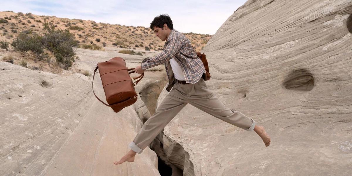 A man in a shirt and trousers leaping barefoot across a crack in pale desert rock, carrying a large suitcase.