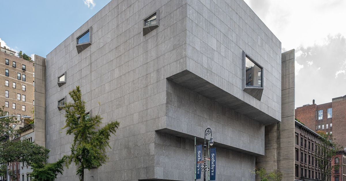 A heavy grey stone building with stepped cantilevered upper floors and small angled windows, on a Manhattan street corner, with banners reading Sotheby's on a lamppost.
