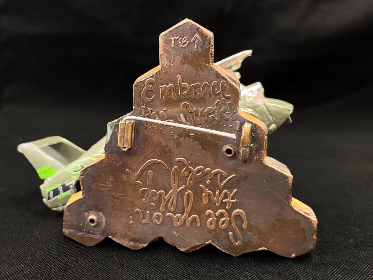 The reverse of the brooch in copper. A clasp sits proud of the surface, and two lines of hand cut lettering run at opposite orientations, one upright and one inverted.