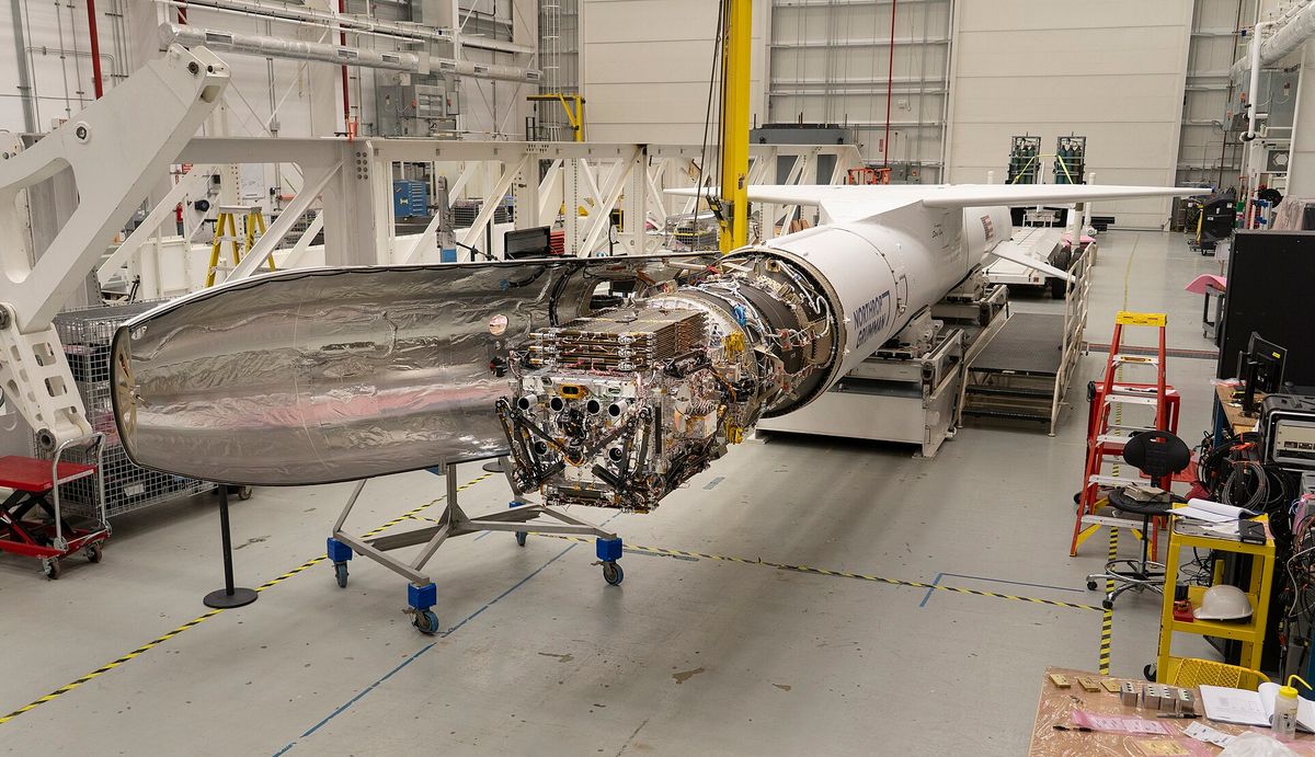 The LINK spacecraft on a trolley in a hangar beside the long white Pegasus XL rocket it will launch on, its metallic surfaces exposed.