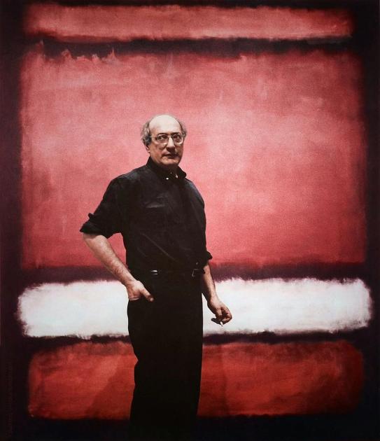 A man in dark clothes standing with one hand on his hip in front of a large canvas of stacked red and white horizontal bands.