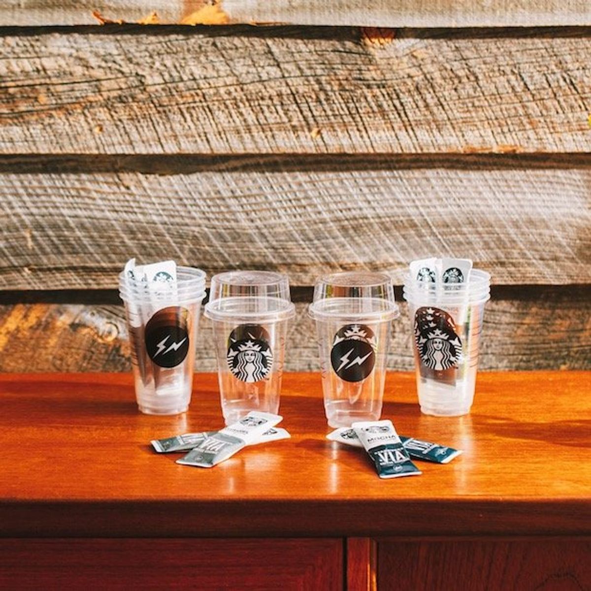 Four clear takeaway cups on a wooden counter, two printed with a lightning bolt roundel and two with a coffee chain logo, sugar sachets in front.