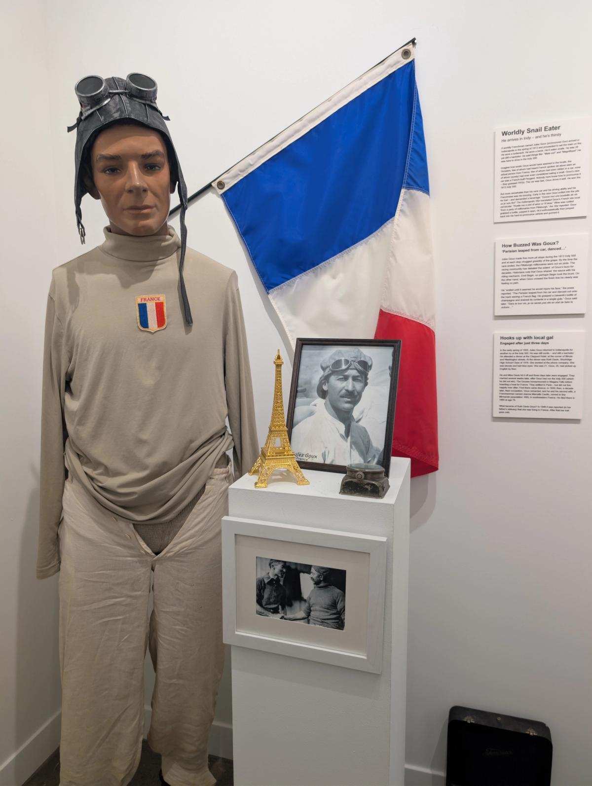 A mannequin dressed in pale racing coveralls and a leather flying helmet with goggles, standing beside a French tricolour, a framed portrait, and a small model of the Eiffel Tower.