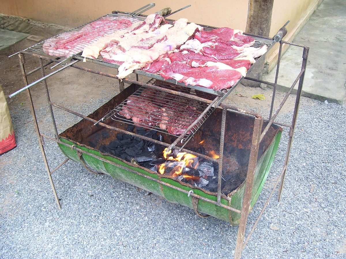 A home-made barbecue welded from reinforcing bar with a green drum firebox, raw cuts of beef laid across the upper rack and charcoal burning below. No people are in the frame.