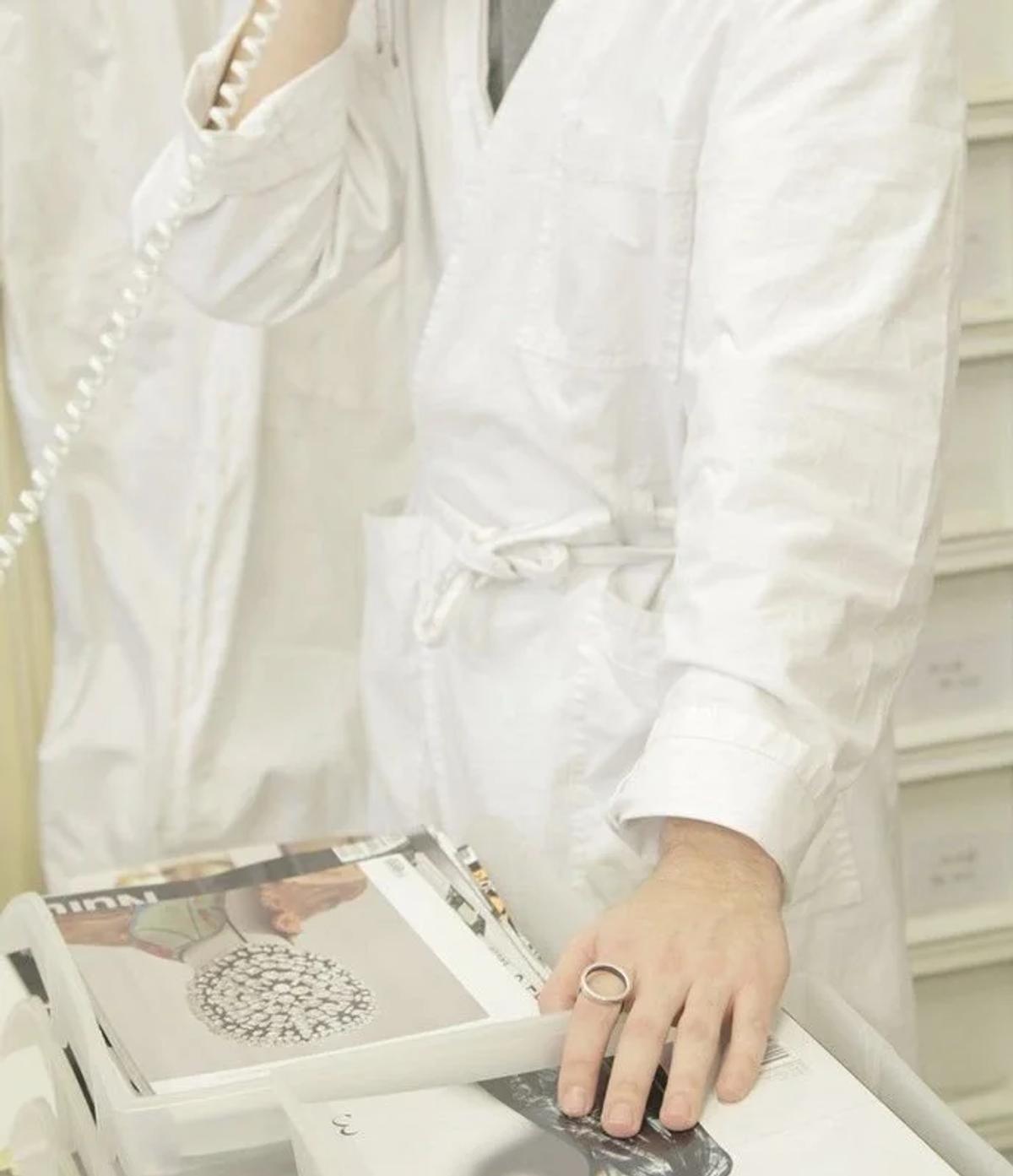 A person in a white cotton coat working at a table of loose prints and magazine pages, one ringed hand resting on the paper.