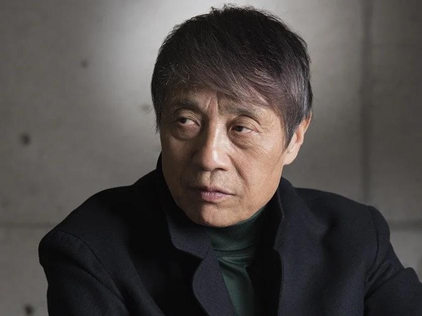 A portrait of an older man with grey hair in a dark jacket and high necked top, photographed against a plain concrete wall.