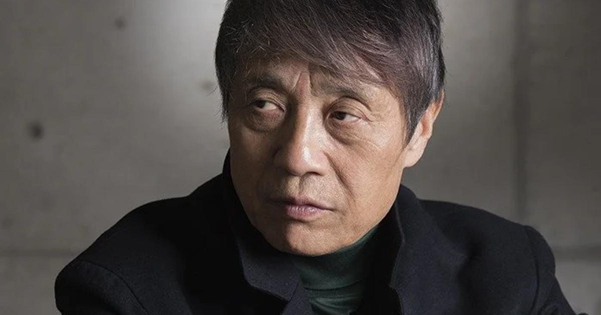 A portrait of an older man with grey hair in a dark jacket and high necked top, photographed against a plain concrete wall.