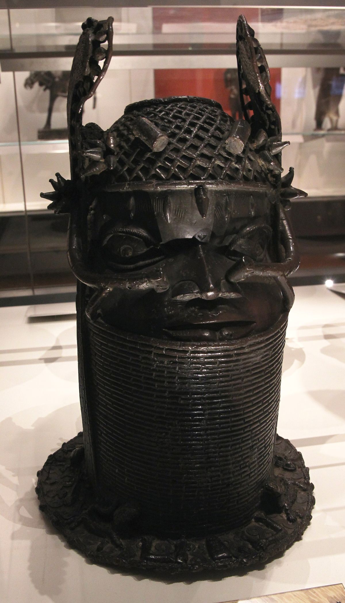A cast brass memorial head of an Oba on a museum plinth, the face framed by a high collar of beaded rings.