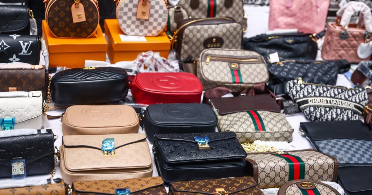 A dense pile of designer handbags, wallets and pouches in assorted monogram and logo prints spread across a table, many still carrying tags.