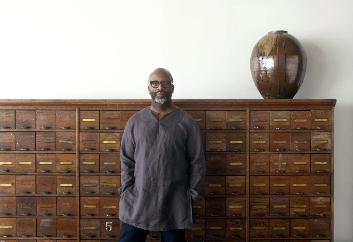 A man in a grey work coat standing in front of a wall of wooden card index drawers, a large glazed ceramic vessel on the ledge above.