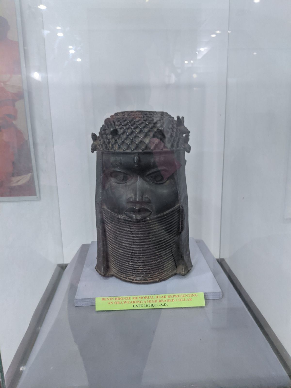 A Benin bronze memorial head inside a glass case, with a hand-typed label on green card giving its description and a late sixteenth century date.