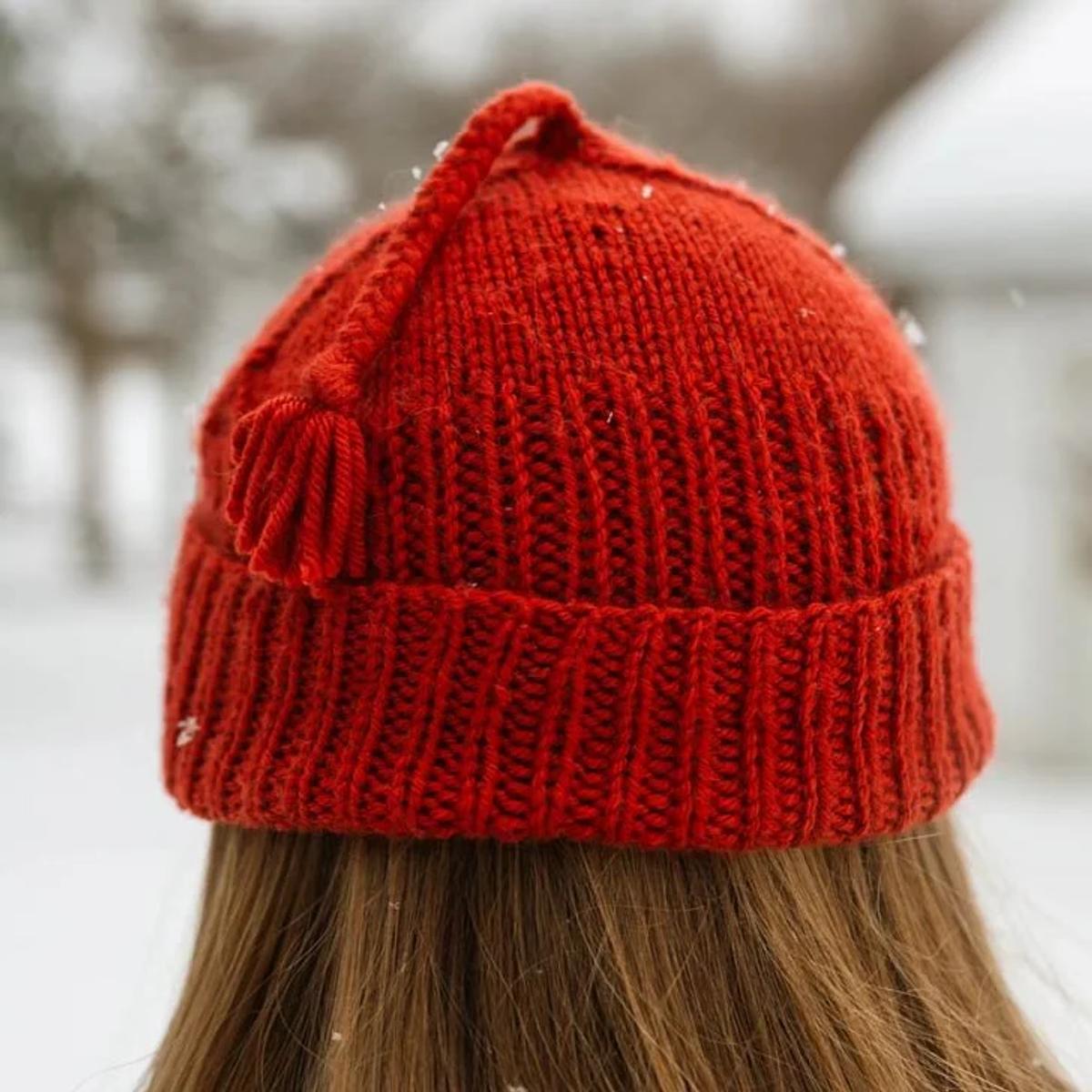 The back of a person's head outdoors in falling snow, wearing a bright red hand knitted ribbed beanie with a tassel.