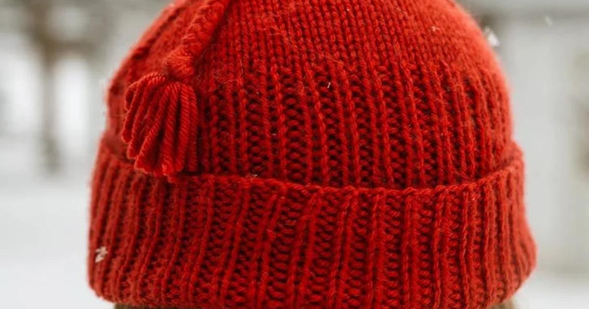 The back of a person's head outdoors in falling snow, wearing a bright red hand knitted ribbed beanie with a tassel stitched to one side.