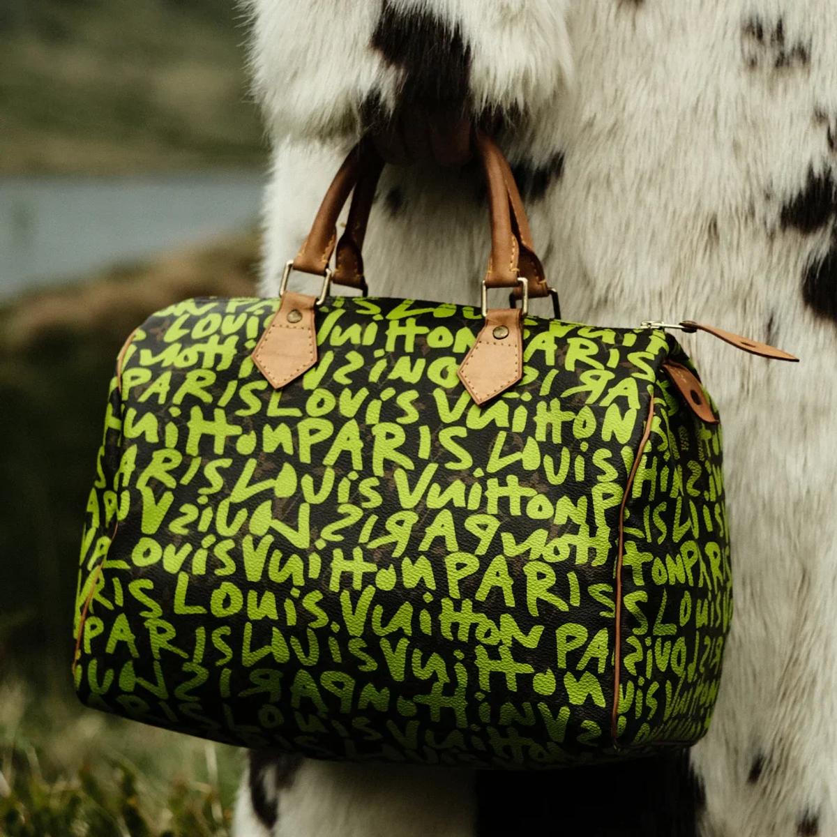 A monogram holdall sprayed with fluorescent green graffiti lettering, held against a black and white cowhide coat outdoors.