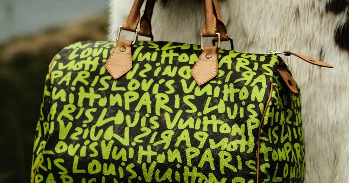A Louis Vuitton Speedy bag in the graffiti print, fluorescent green lettering sprayed across dark brown canvas, held against a black and white cowhide coat outdoors.
