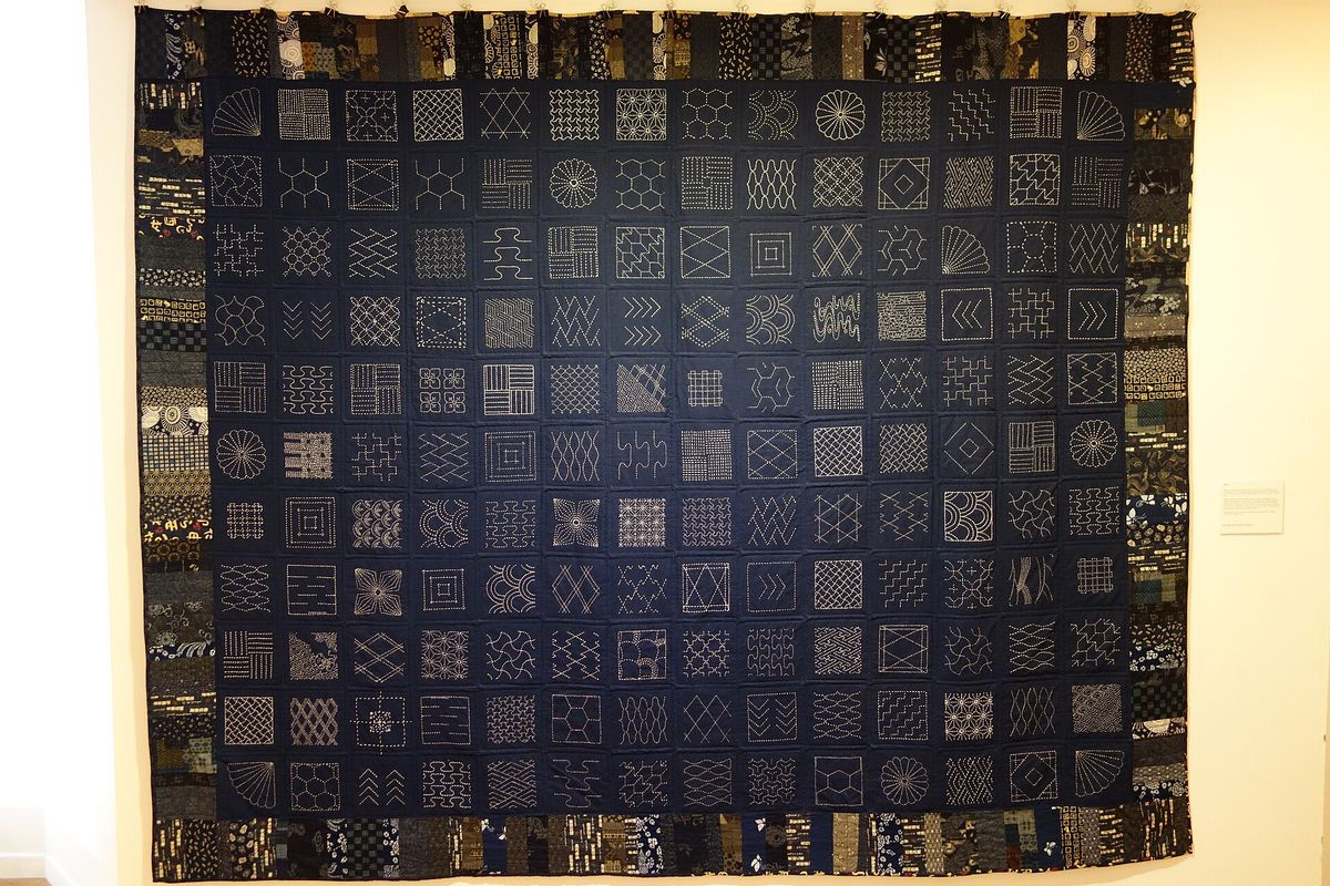 A large indigo hanging in a gallery, divided into around a hundred and fifty squares, each stitched with a different white sashiko pattern, bordered with patterned cloth.