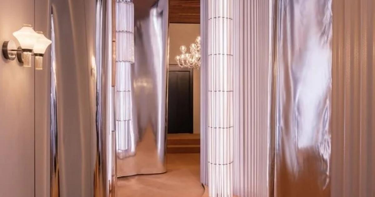 A narrow corridor lined with curved polished metal panels and tall fluted glass light columns, a sand coloured carpet underfoot and a chandelier visible at the far end.