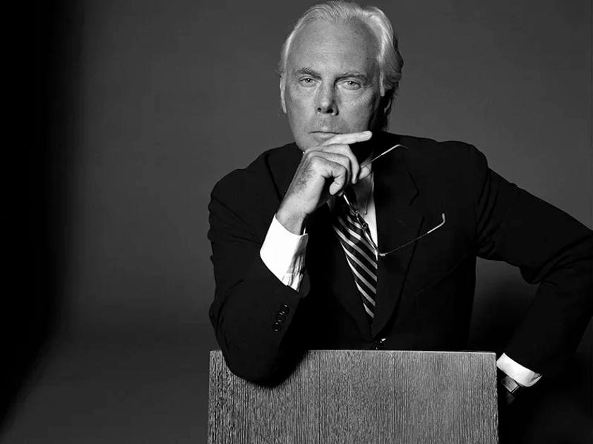 A black and white portrait of Giorgio Armani in a dark suit and striped tie, seated with one hand raised to his chin, holding his glasses.