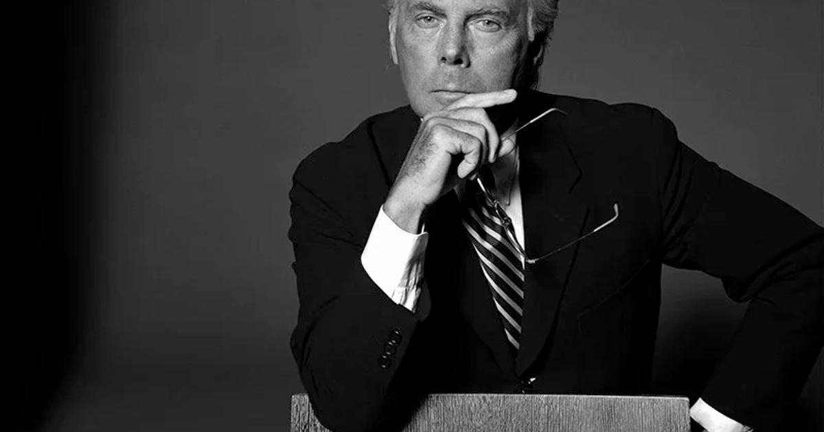 A black and white portrait of Giorgio Armani in a dark suit and striped tie, seated with one hand raised to his chin, holding his glasses.