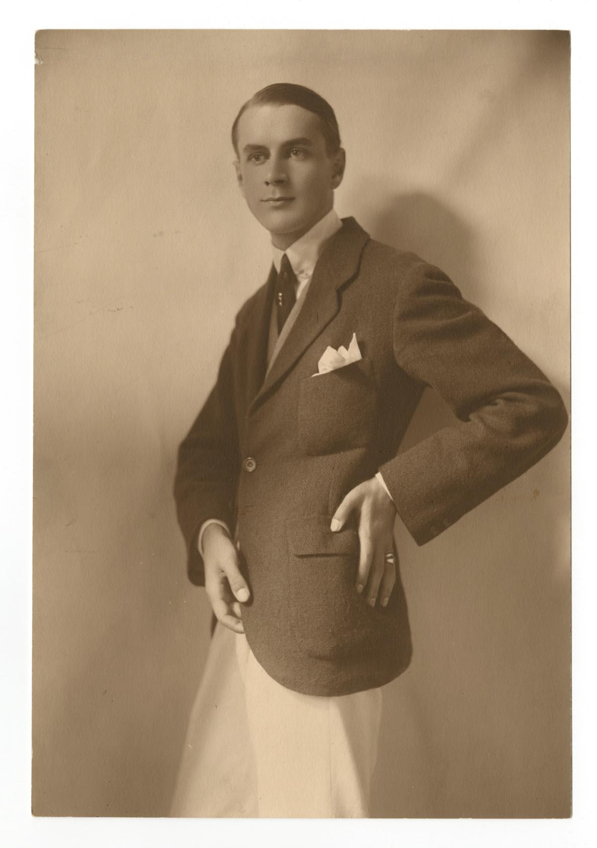 Sepia studio photograph of a young man in a tweed jacket and pale trousers, hand on hip, pocket square showing.