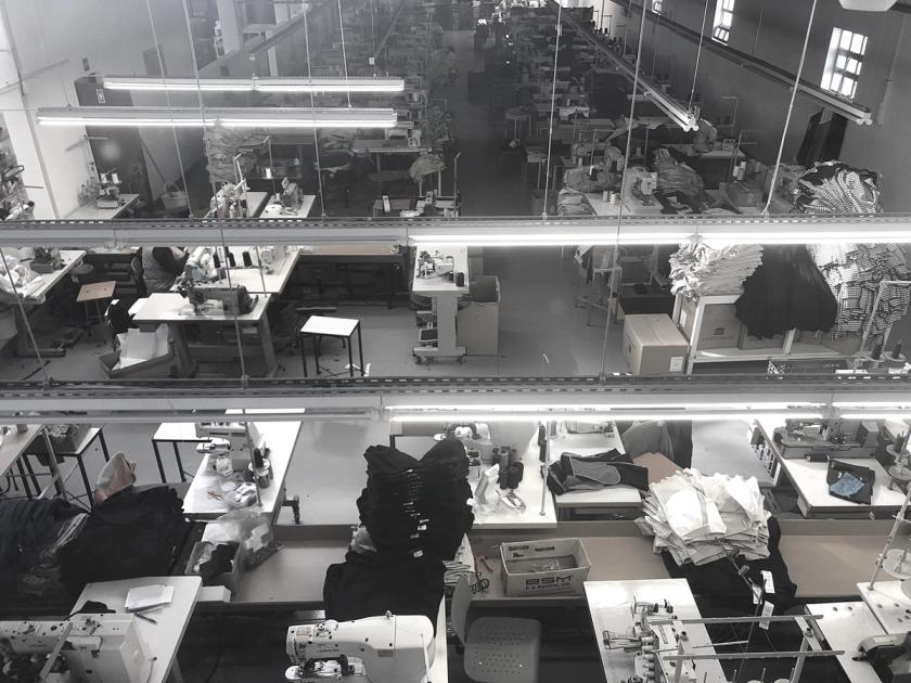 A garment factory floor in Portugal seen from above, rows of sewing stations under strip lights with stacks of identical cut pieces beside each machine.