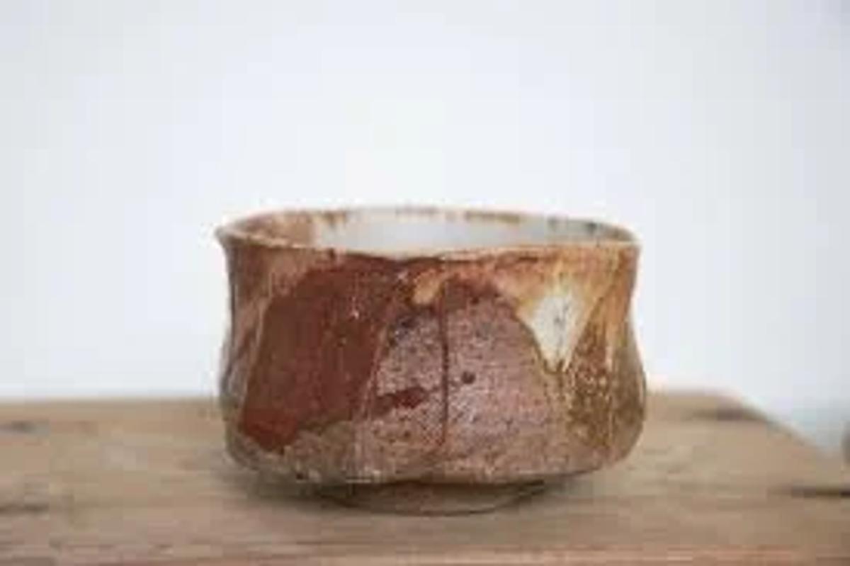 A small hand thrown tea bowl with a rough foot and uneven rim, glazed in patches of rust brown and cream, standing on a wooden board.