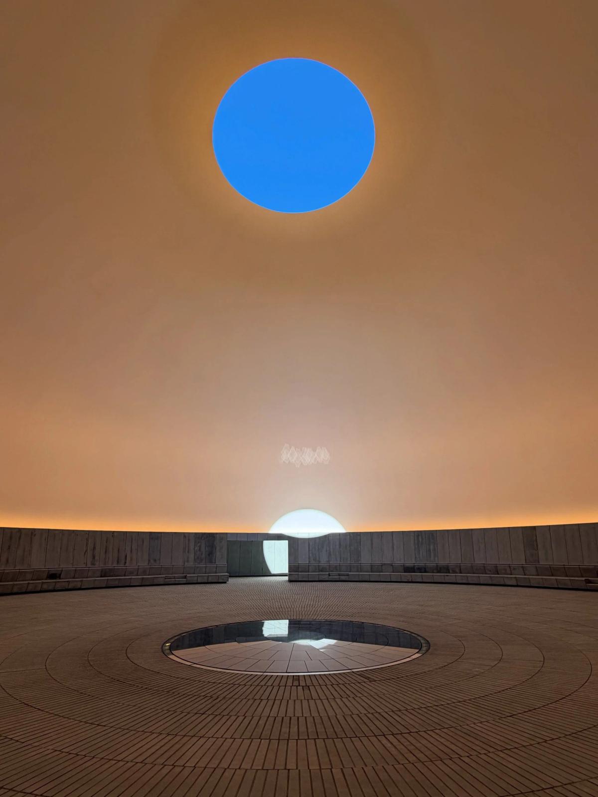 A circular chamber lit warm gold with an oval opening cut through the ceiling to a deep blue sky, a low pool at the centre of the floor.
