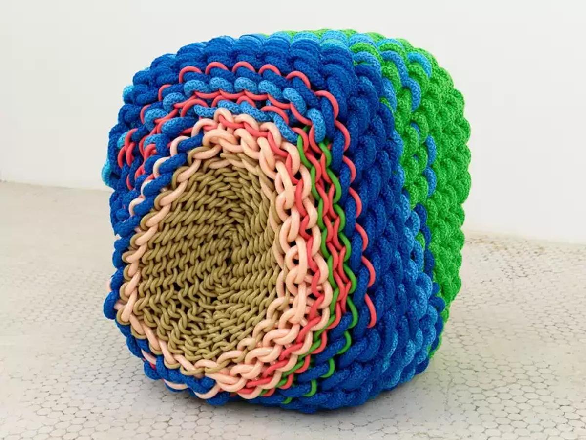 A cube shaped object built from thick coiled rope in concentric rings of blue, green, red, cream and olive, resting on a tiled floor.