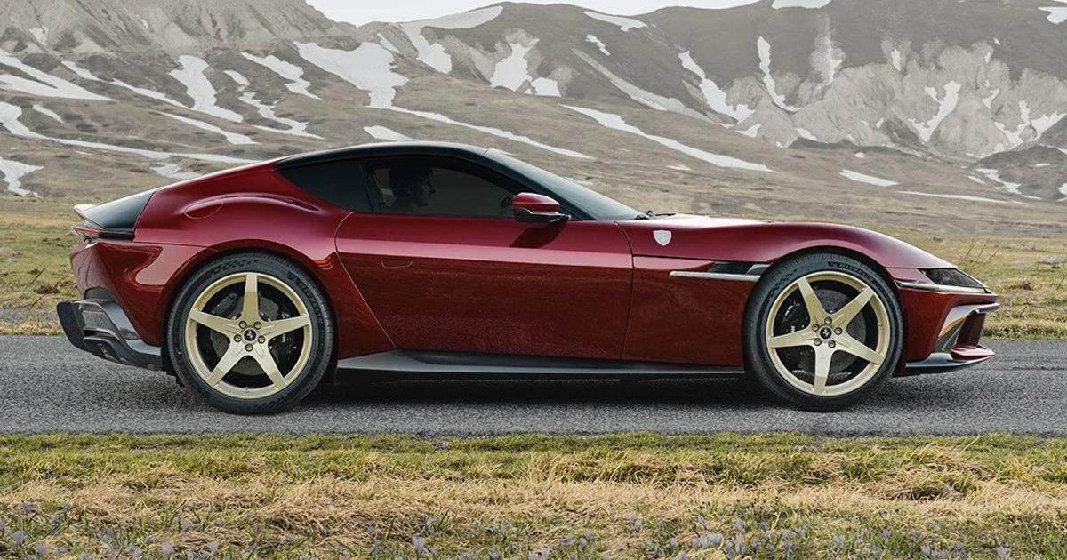 A dark red Ferrari 12Cilindri photographed in profile on an empty mountain road, gold five spoke wheels, snow streaked peaks behind and small purple crocus in the grass.