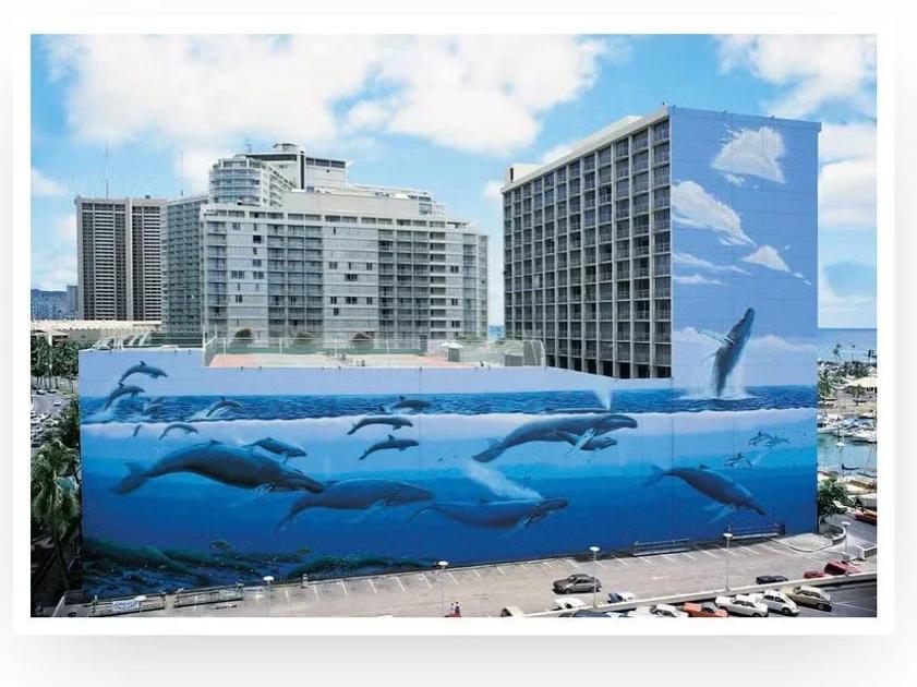A long ocean mural of whales and dolphins swimming in blue water, painted across the base of a tall city building beside a coastal road and car park.
