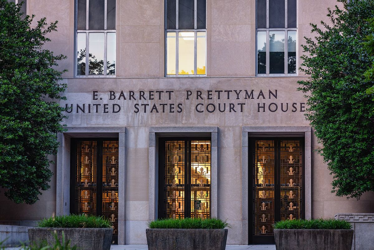 The lettered stone facade and lit bronze doors of the E. Barrett Prettyman United States Court House in Washington, framed by trees. No people are in the frame.