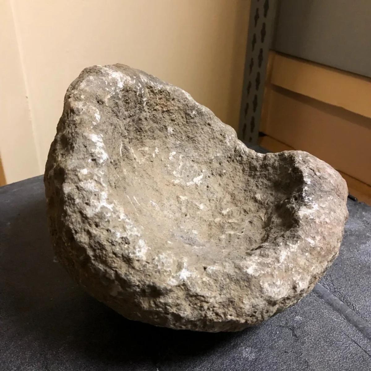 A rough hewn stone basin with a deep hollowed bowl and an uneven broken rim, standing on a grey floor against a plain wall.