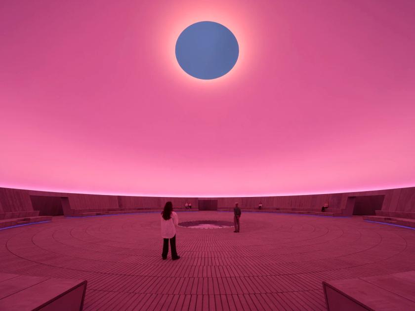 A circular chamber lit deep pink with an oval opening cut through the ceiling to blue sky, two small figures standing on the patterned floor below.