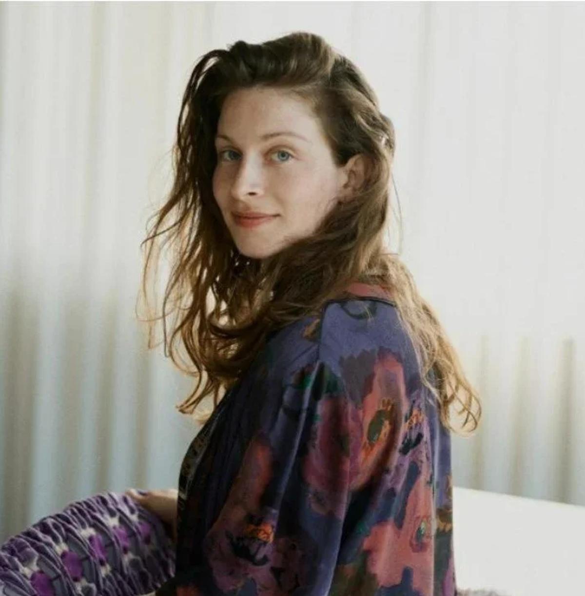 A portrait of a woman with long wavy hair turning to the camera, wearing a printed silk robe against a pale curtain.
