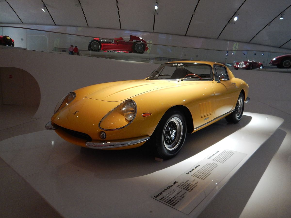 A yellow Ferrari 275 GTB/4 displayed in a museum interior with a curved white ceiling above it.