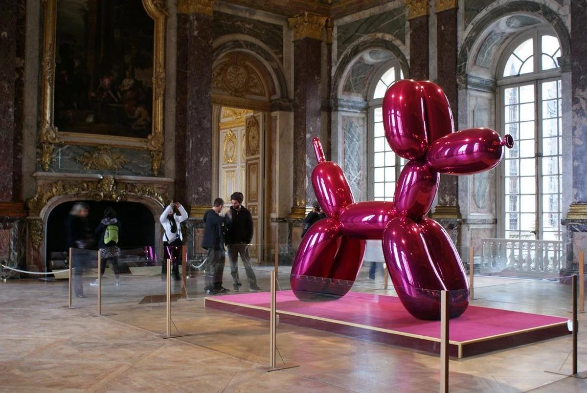 A large mirror polished magenta balloon dog sculpture on a pink plinth in a gilded palace room with tall windows and visitors around it.