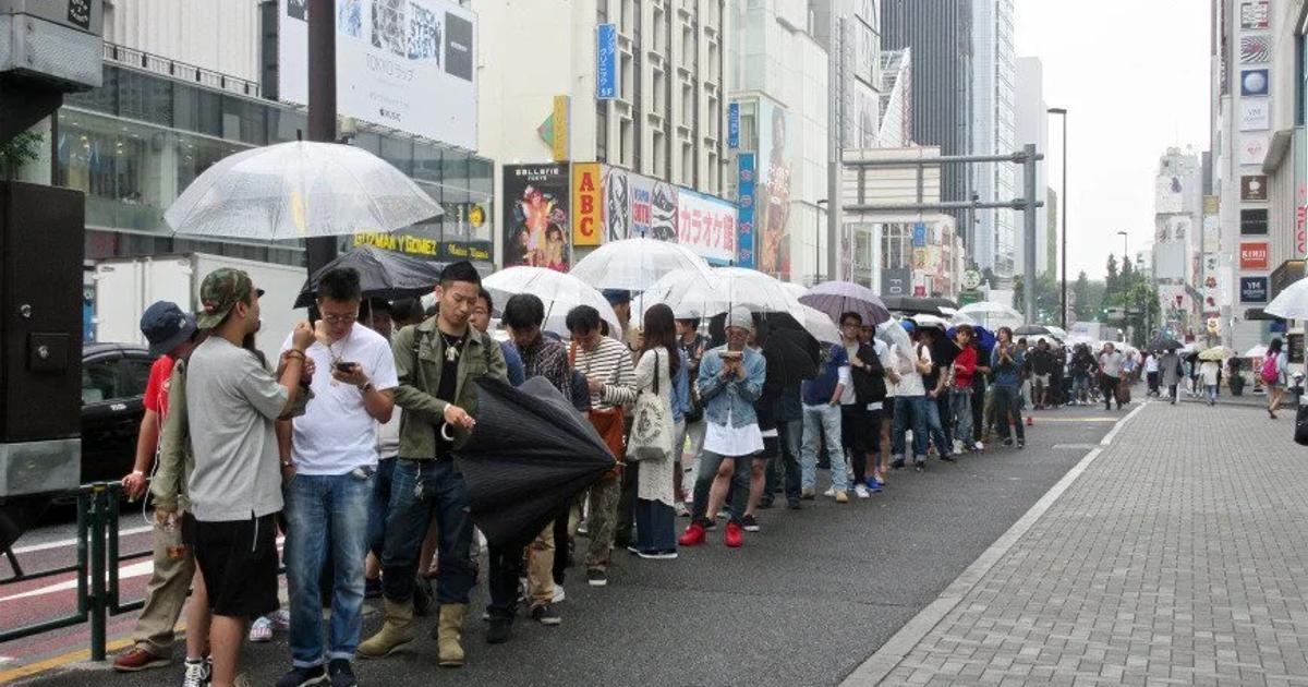 A long queue of people with clear umbrellas waiting along a wet city pavement in Japan, shopfronts and vertical signage rising behind them.